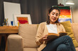 © Chanakon - Asian beautiful woman sitting reading book on cozey couch sofa in living room with warm light at night. People female stay home part time relax at home.