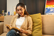© Chanakon - Asian beautiful woman sitting reading book on cozey couch sofa in living room with warm light at night. People female stay home part time relax at home.