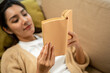 © Chanakon - Asian beautiful woman lay down on cozey couch sofa reading book in living room with sun light morning. People female stay home part time relax at home.