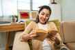 © Chanakon - Asian beautiful woman sitting reading book on cozey couch sofa in living room with sun light morning. People female stay home part time relax at home.