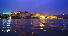 Udaipur City Palace On The Bank Of Beautiful Lake Pichola At Sunset, Rajput Architecture Of Mewar Dynasty Rulers Of Rajasthan. Water Ripples Reflect Night City View. Udaipur, India, Asia