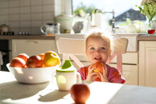 Happy Girl Holding Apple Fruit Sitting At Dining Table In Kitchen