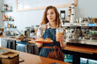 © maxbelchenko - Smiling woman barista works in a cafe. A young woman in an apron behind the bar holds to-go coffee and desserts for ordering.