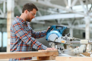 Wall Mural - A carpenter works on woodworking the machine tool. Man collects furniture boxes. Saws furniture details with a circular saw. Process of sawing parts in parts. Against the background of the workshop.