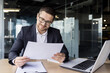 © Tetiana - A happy young businessman is sitting in the office at the desk and looking at the documents he is holding in his hands