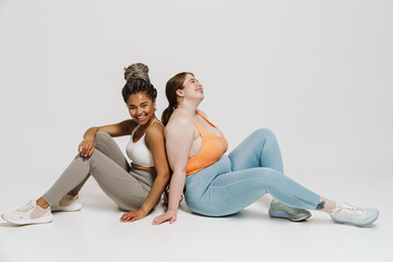 Wall Mural - Two happy women posing together while sitting isolated over white wall