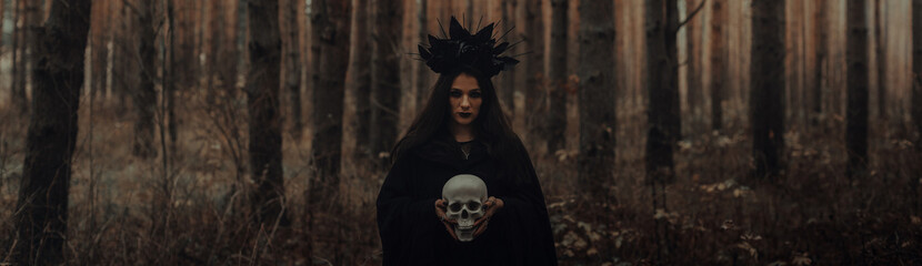 black terrible witch with a skull in the hands of a dead man in the woods. Panoramic wide horizontal photo for banner head cover