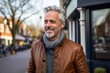Portrait of a handsome middle-aged man with grey hair and beard wearing a brown leather jacket on a city street