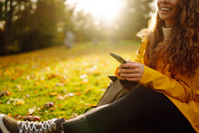 A Smiling Woman Sits Outdoors On The Lawn Among Yellow Autumn Leaves With A Phone In Her Hands. A Female Uses A Smartphone In An Autumn Park, Enjoying Nature.