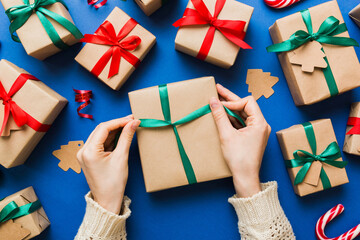 Woman is packing surprise gift for Christmas holidays, sitting at workplace with craft tools, top view. Woman wrapping Christmas gift at colored table