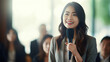 © Santy Hong - A young, confident Asian corporate businesswoman, smiling as she holds a microphone, delivering a candid presentation in a boardroom filled with attentive colleagues.