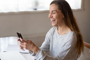 Wall Mural - Excited laughing woman looking at phone screen, having fun, holding smartphone in hands, making video call, chatting online, sitting at table, posing for selfie, browsing mobile device apps