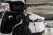 African american female with cell phone inserting charger gun into electric car port in bottom-level garage. Effective employee in business outfit using parking time for EV fill-up and communication.