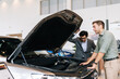 © dikushin - Friendly car dealer describing engine to doubtful male buyer in showroom talking gesturing standing near automobile with open motor hood. Concept of choosing and buying new auto in dealership