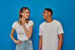 © LIGHTFIELD STUDIOS - cheerful woman posing with hand in pocket and looking at african american friend on blue background