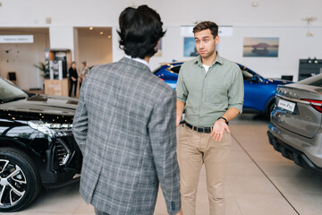 Wall Mural - Rear view of unrecognizable professional male car seller with serious male customer in auto dealership, talking about specifications and performances of vehicle. Customers looking at cars in showroom.