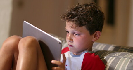 Wall Mural - Young boy holding tablet device at night. Child kid staring at screen playing video-game online