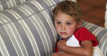 Wall Mural - Young boy toddler watching TV at home lying on sofa. Child watches content on couch in the evening