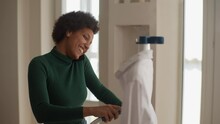 Cheerful African-American Young Housewife Talking Smartphone And Steaming White Shirt With Manual Garment Steamer At Home. Happy Black Female Using Steam System For Ironing Clothes At House.