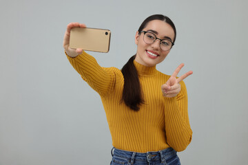 Canvas Print - Smiling young woman taking selfie with smartphone and showing peace sign on grey background