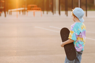 Fashionable girl in casual clothes and jeans holds a skateboard in her hands, back view with copy space. The concept of children's street fashion and outdoor activities.