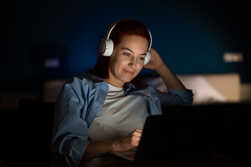 woman with laptop watches video with headphones. calm girl happily smiles quite broadly using comput