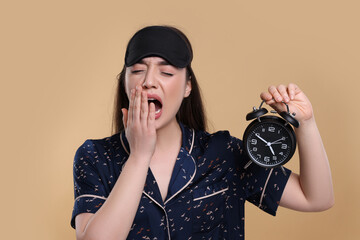 Canvas Print - Tired young woman with sleep mask and alarm clock yawning on beige background. Insomnia problem