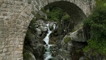 Flying Under Arch Of Brick Bridge While River Water Stream Is Flowing In Summer Season In Furka Pass Valley Of Switzerland. Aerial Drone View