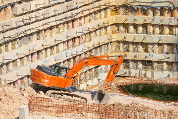 Wall Mural - Pit of a metro station with piles ground, working orange excavator dig and extract soil. Excavator stands on a slope descending into a quarry pit. Puddle of groundwater