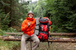 male tourist with backpack and hiking equipment sits resting in the forest, guy in orange jacket on mountain