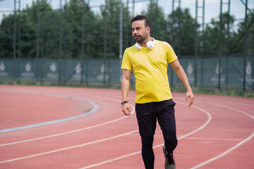 Poster - Bearded man in yellow t-shirt with wireless headphones standing on empty running track with red surface and white marking at contemporary sports ground in urban park
