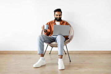 Wall Mural - Arab Guy Using Laptop Holding Cup Of Coffee Sitting Indoor