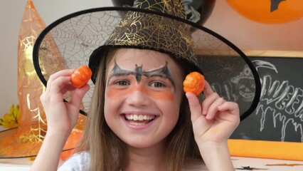 kid girl in black hat smiling playing with little toy halloween pumpkins at home on background of de