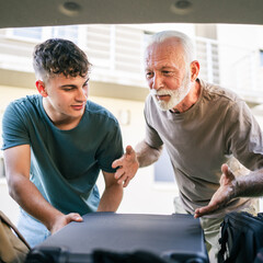 Wall Mural - Two men Teenager and senior grandfather pack baggage in trunk of car