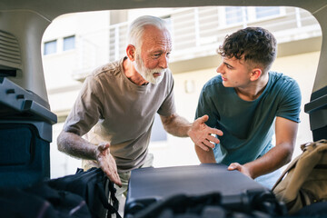 Wall Mural - Two men Teenager and senior grandfather pack baggage in trunk of car