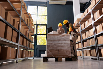 Diverse employees wearing industrial overall and helmet working in warehouse, preparing clients orders for delivery. Storage room workers checking shipping detalis on laptop computer.