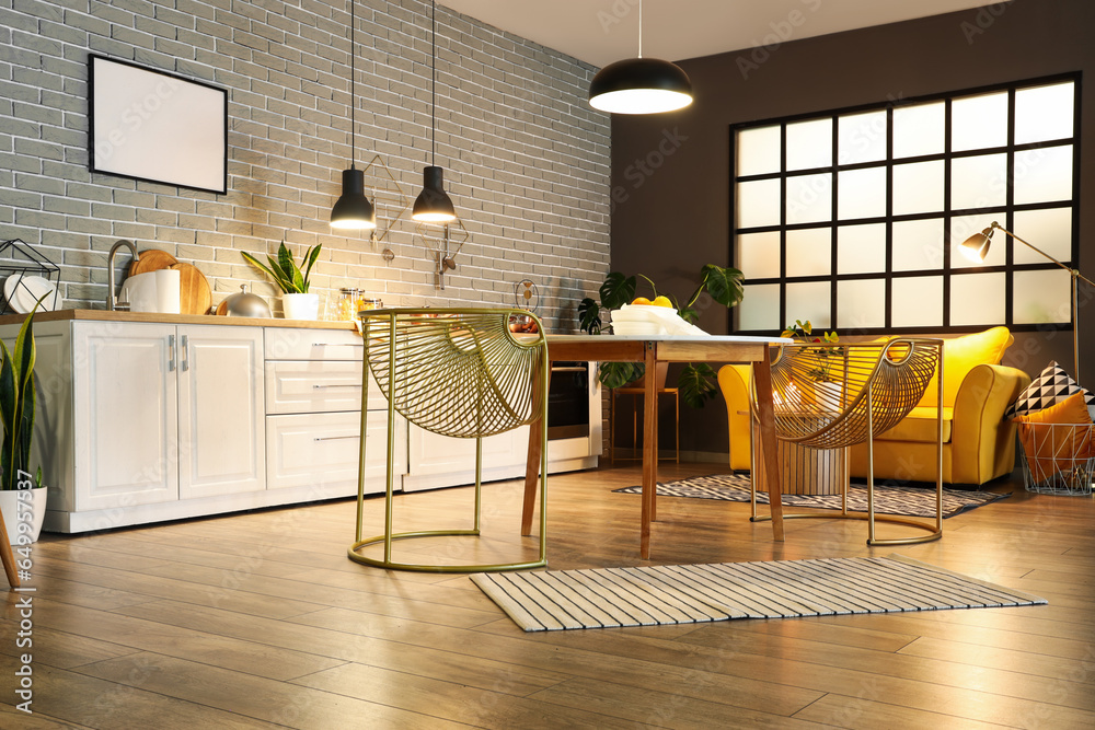 Interior of modern kitchen with glowing lamps, dining table, yellow sofa and white counters at evening