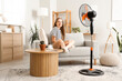 © Pixel-Shot - Young woman with mobile phone and blowing electric fan at home