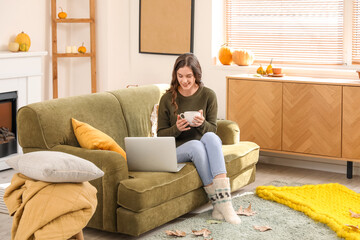 Wall Mural - Young woman with cup of tea and laptop sitting at home on autumn day