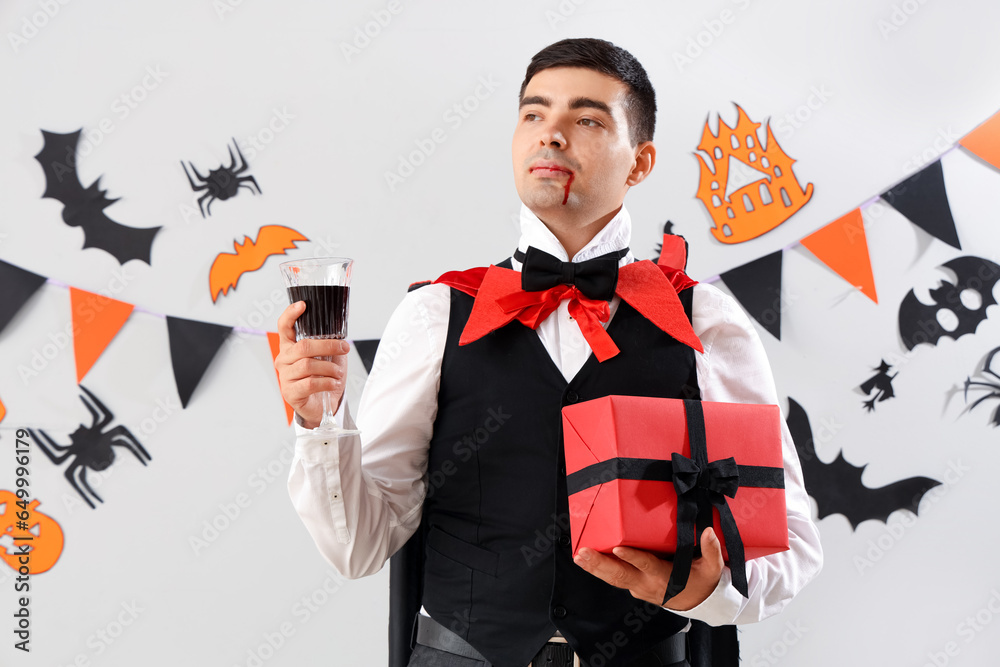 Young man dressed for Halloween as vampire with gift and drink on light background