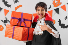 Little Boy Dressed For Halloween As Vampire Pointing At Gifts On Light Background