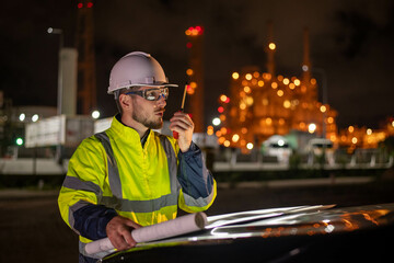 Engineers wear uniform and helmet stand in front of the car hand holding blueprint paper, survey inspection work plant site use radio communication to work with night lights oil refinery background.