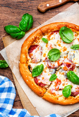 Wall Mural - Pizza with italian salami sausage, mozzarella cheese, tomato sauce and green basil on rustic wooden kitchen table background, top view