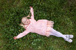 © somemeans - Little blonde girl in pink dress and jacket is lying on green grass with her arms outstretched to the sides. Top view.
