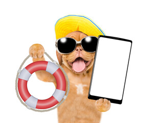 Poster - Happy mastiff puppy wearing summer hat and sunglasses shows lifebuoy and holds big smartphone with white blank screen in it paw. Isolated on white background