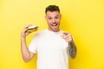 Wall Mural - Young caucasian man holding a burger isolated on yellow background surprised and pointing front