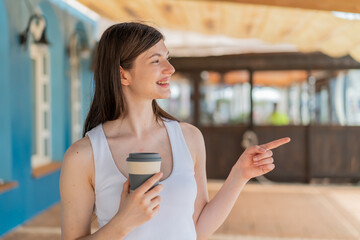 Wall Mural - Young pretty Ukrainian woman holding a take away coffee at outdoors pointing to the side to present a product