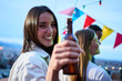 © CarlosBarquero - Portrait pretty young Caucasian woman toasting bottle beer in hand smiling. Attractive girl posing cheerful for photo at party friends outdoor at sunset. People gathered on rooftop celebrating success
