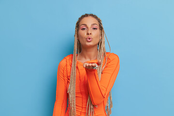 Wall Mural - Kind blonde woman dressed in orange long sleeve top sends air kiss while standing on blue background in studio, lucky day concept, copy space
