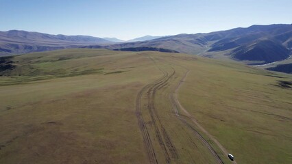 Wall Mural - A white SUV is driving through mountain green fields. In the distance there are high peaks with snowy peaks. Dirty road. Blue sky. A dog runs and shepherd rides. Mountainous terrain, sometimes forest